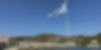 The Greek flag waving proudly on a pole at the Paxos harbor, with a clear blue sky overhead and traditional buildings and boats in the distance, capturing the patriotic spirit of the island